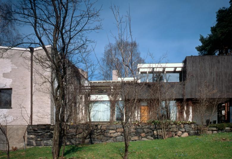 Kuvassa on Aaltojen kotitalo, joka on modernin näköinen ja siinä on kaksi siipeä.  Talo on ällän  muotoinen ja siinä on isot ikkunat.  Talon ympärillä on lehtipuita.