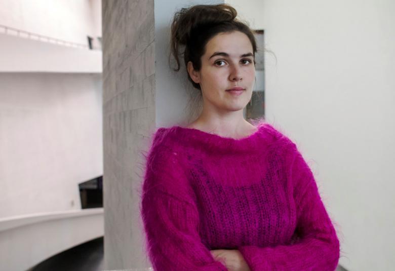 Kuvassa on Emmi Jääskeläinen, joka nojaa valkoiseen hyllyyn.  Hänestä on puolivartalokuva ja hänellä on lilan värinen villapusero.