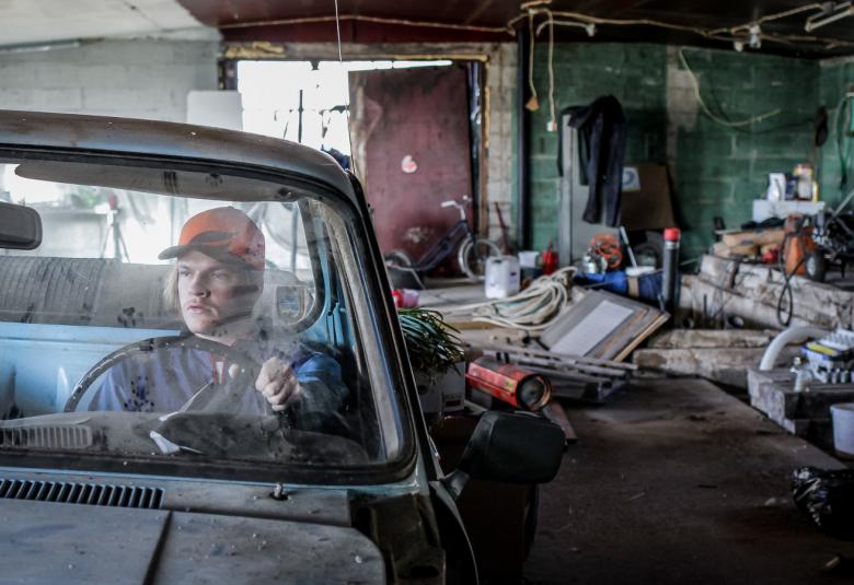 Kuvassa on Juho Uusitalo auton etuistuimella ja auto on autokorjaamolla.  Taustalla näkyy erilaista autokorjaamotavaraa.