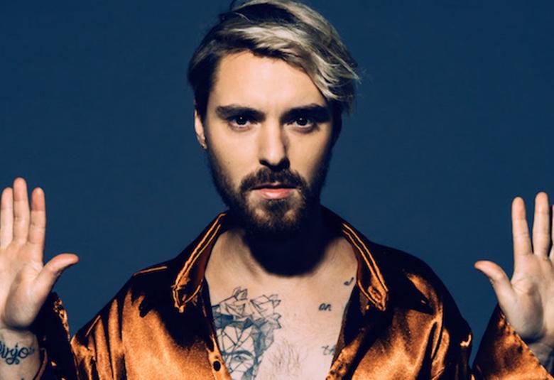 Kuvassa on LEO, jolla on molemmat kädet ylhäällä.  Hänellä on kuparin kiiltävä paita  päällä ja avoimista napeista näkyy tatuointia. 