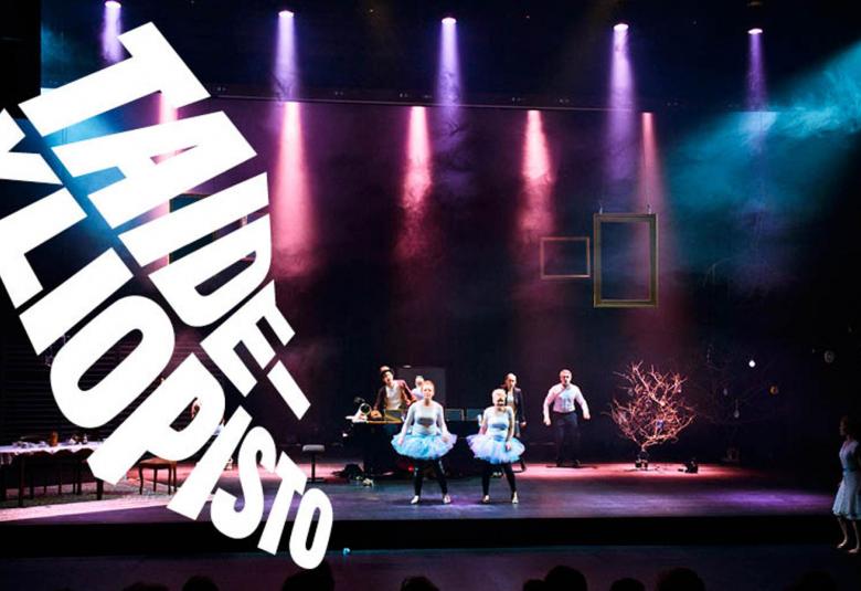 Kuvassa on valkoisella Taideyliopiston logo vinosti vasemmalla.  Kuvassa on näyttämöesitys pimennetyssä katsomossa ja valaistulla näyttämöllä.