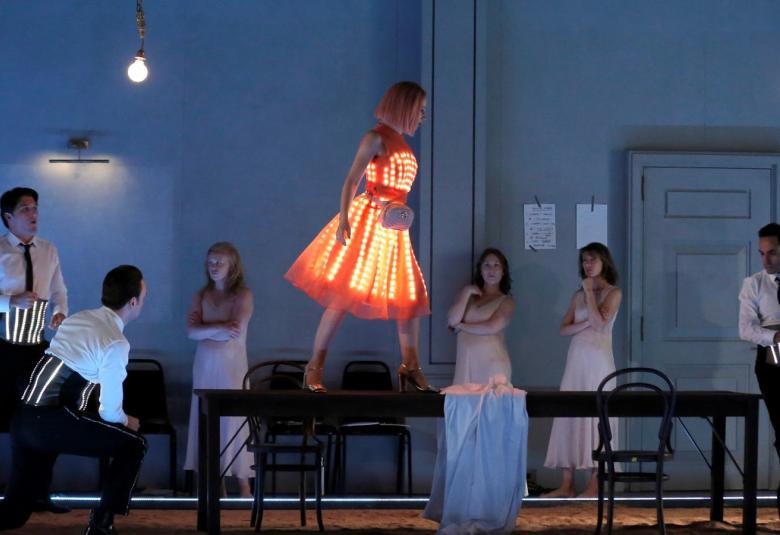 Kuvassa on nuori nainen pöydän päällä oranssisessa pallollisessa mekossa.  Pöydän ympärillä on sekä miehiä että naisia ja heilla on siistit vaatteet päällään.