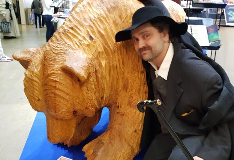 Kuvassa on Eino Leino, joka nojaa puiseen isoon karhuveistokseen.  Eino Leino on pukeutunut mustaan pukuun, viittaan ja lierihattuun ja on kyykistyneenä karhun viereen.