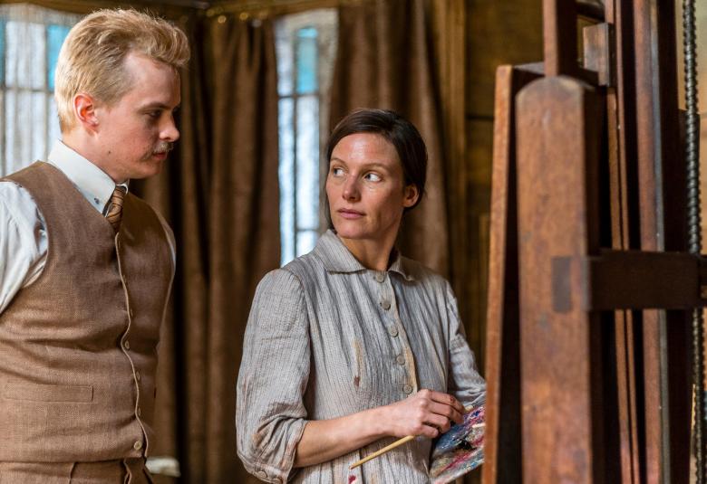 Kuvassa ovat Johannes Holopainen ja Laura Birn seiromassa taiteilija-ateljeessa, joka on ruskean sävyinen. Huone on puukalusteinen.  