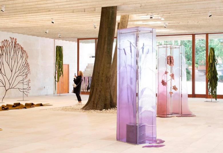 Kuvassa on Sääennuste tiulevaisuudelle -näyttelyä Venetsian biennaalessa.  Kuvassa on vaalealla lattiapinnalla on huputettuja isoja huonekasveja, puunrunkoa ja seinällä iso kaksiulotteinen pensas.