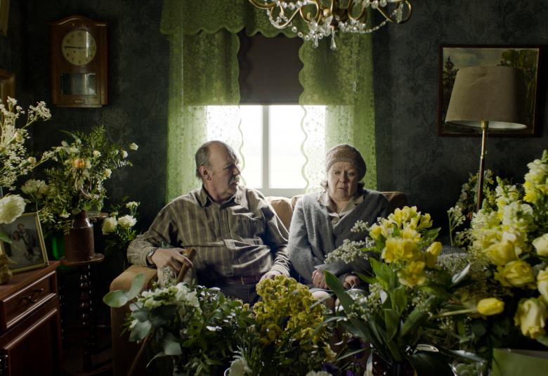 Kuvassa on Peik Stenberg ja Lena Labart istumassa sohvalla ja edessä sekä ympärillä on kukkasia.