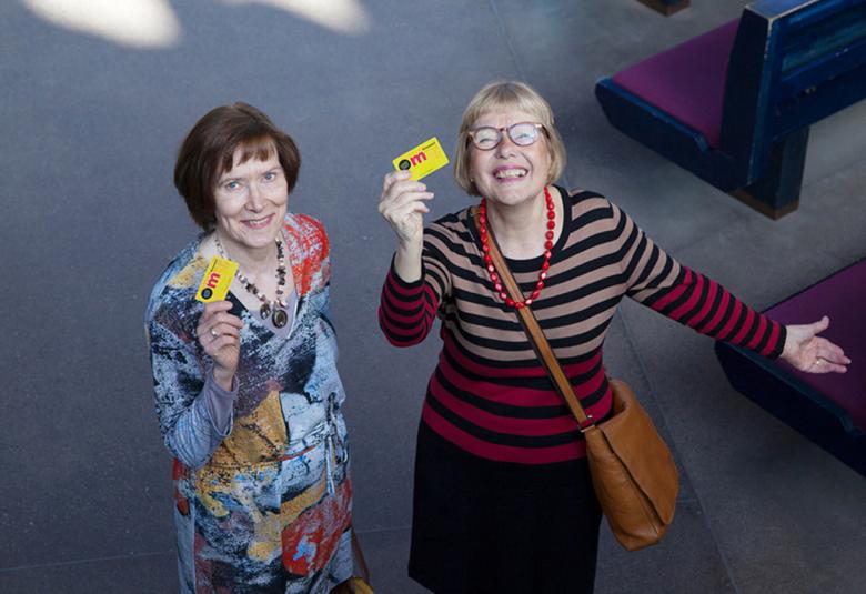 Kuvassa on kaksi keski-ikäistä naista kuvattuna ylhäältä päin.  Oikealla puolella olevalla on oikeassa kädessään museokortti, jota hän iloisesti näyttää. 