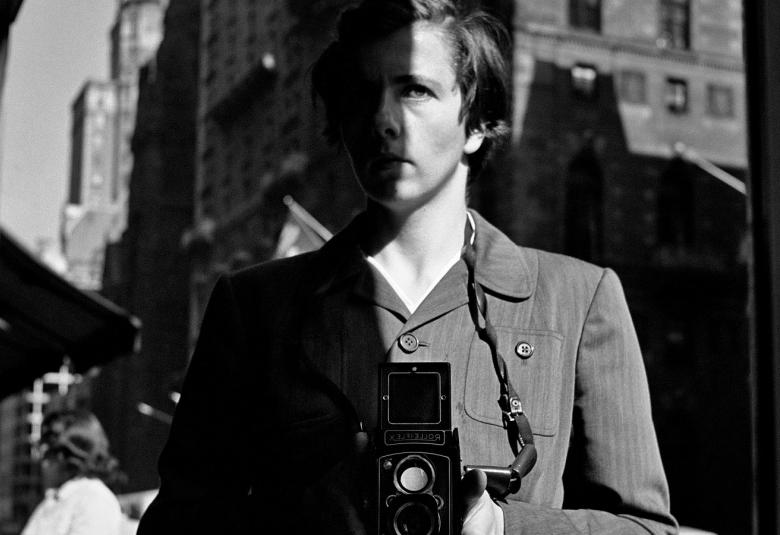 Kuvassa on Vivian Maier kamera kädessään vyötärön alapuolella.  Kuva on otettu New Yorkissa kadulla ja se on msuta-valkoinen.