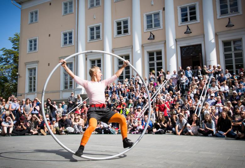 Kuvassa on Tampereen teatterikesän esitys vaalean keltaisen rakennuksen edessä, jossa on yleisöä. Keskellä on esitys, jossa on mieshenkilö ison vanteen sisällä. Sää on lämpimän aurinkoinen.
