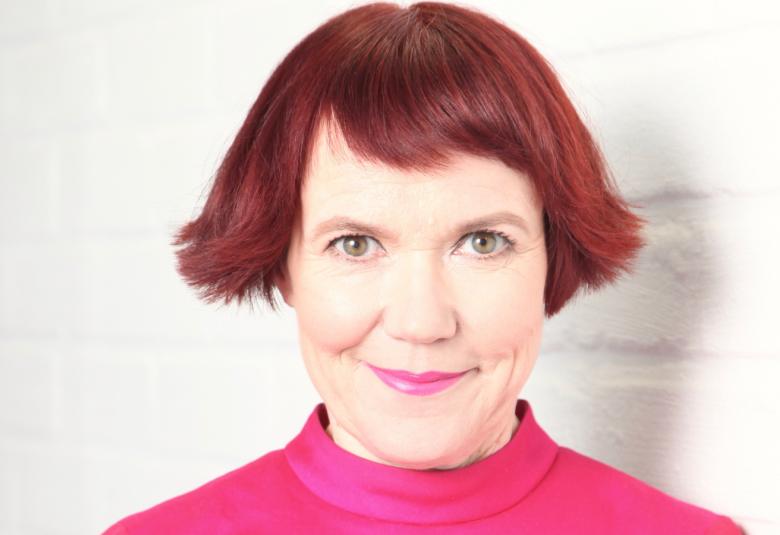 Kuvassa on Rosa Liksom ja hänellä on punainen polkkatukka sekä pinkki pusero.  Kuva on kasvokuva.