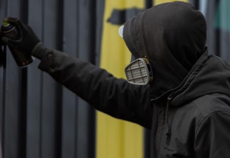 Kuvassa on hupparissa oleva henkilö, joka käsittelee spraylla seinässä olevaa verhoa.  Verhot ovat eri värisiä.  