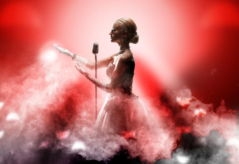 Kuvassa on Helena Rängman punaisessa tyllihelmaisessa puvussa mikrofoni edessään. Kuvassa on valkoisen vaaleanpunaista höyryä melkein vyötärölle asti.