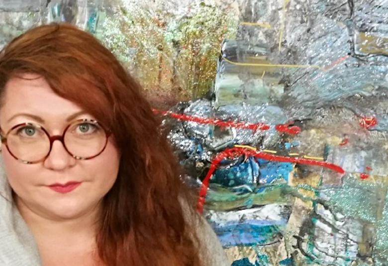 Kuvassa on Minttu Saarinen vasemmalla ja takana on taulu Strong wind, joka abstrakti siniharmaa -sävyinen.  Keskellä on punainen juova.