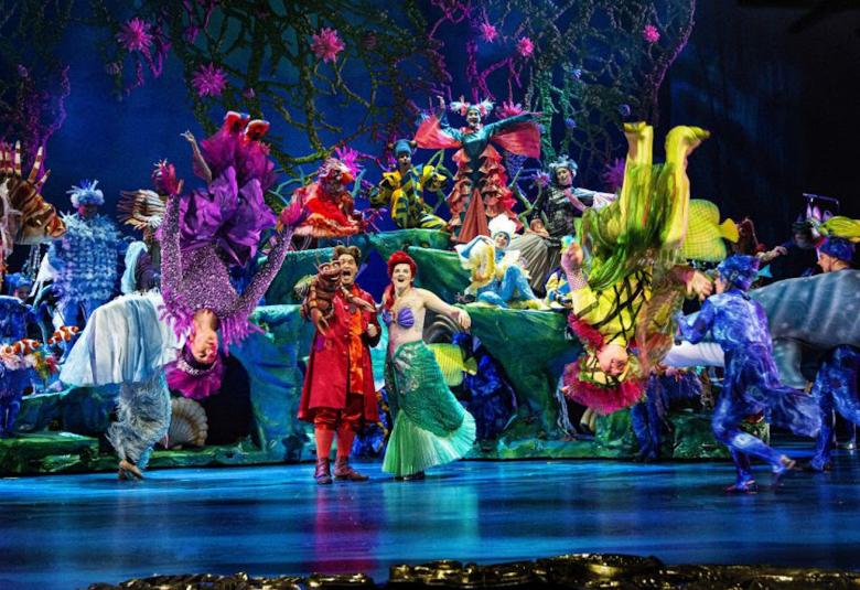Kuvassa on värikkäitä tanssijoita etualalla ja laulajat keskellä.  Taustalla on värikäs lavastus ja siinä värikkäästi pukeutuneita olentoja kulissien eri tasoilla. Lattia on syvän sininen.
