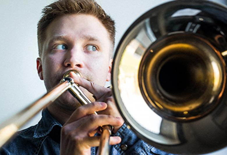 Kuvassa on trumpetisti ja kuva on otettu suoraan edestä. Trumpetti on etualalla ja soittaja on mies.  Kuva on rajattu pelkästään trumpettiin ja soittajan kasvoihin.