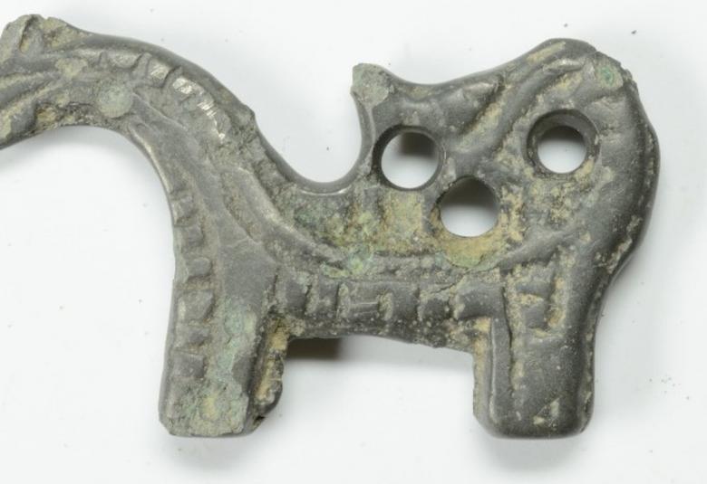 Kuvassa on eläinhahmoinen riipus, jolla on kuono ja takana kolme reikää.  Väri on harmaa ja siinä on vihertäviä laikkuja.