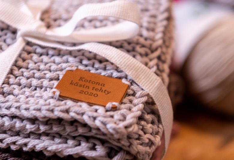 Kuvassa on kudottua ruskeaa neuletta ja se on paketoitu vaalean sinisellä nauhalla.  Neuleessa on merkki nahkainen Kotona käsin tehty 2020.