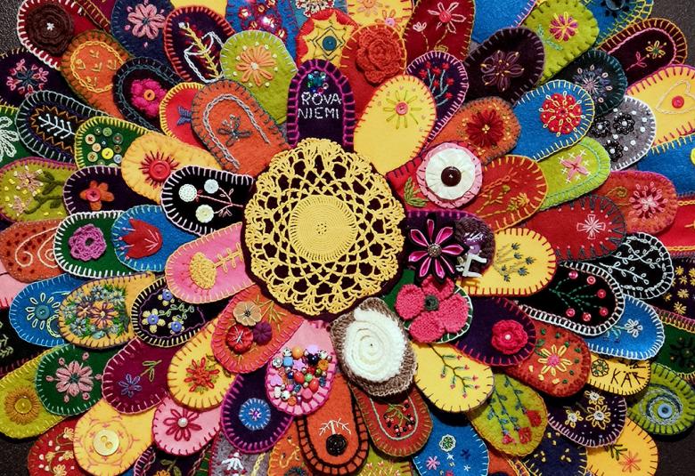 Kuvassa on kukkamaisessa asetelmassa iso matto, jossa on värikkäitä lehtimäisiä virkattuja kukkamaton osia.