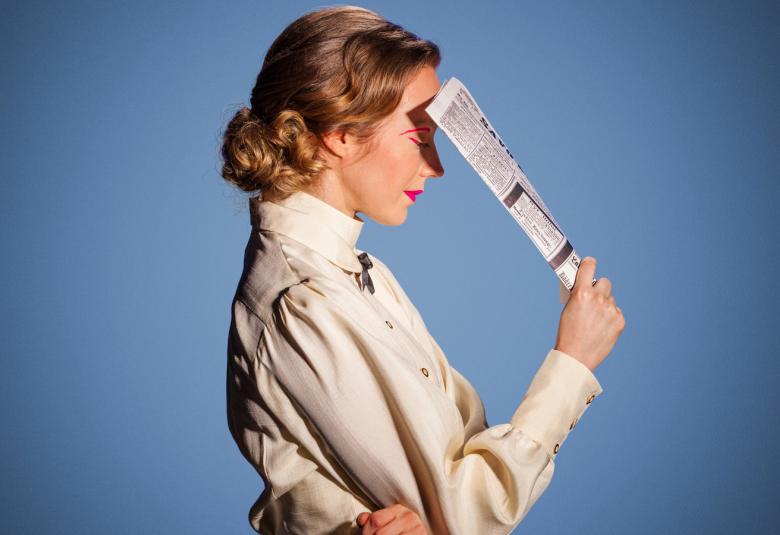 Kuvassa on Minna Canthia esittävä näyttelijä Heidi Ajanto ja hänestä on sivukuva.  Hänellä on sanomalehti kädessään ja nojaa sillä kumartuneeseen päähänsä.