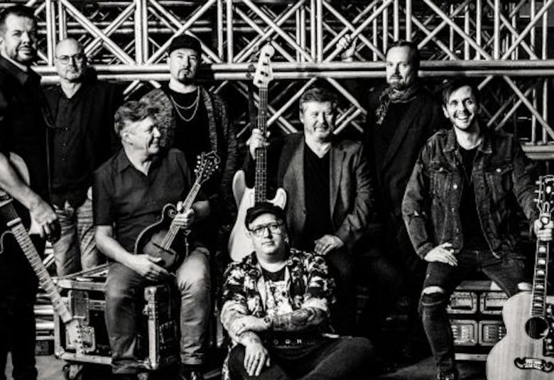 Kuvassa on Arttu Wiskari istumassa lattialla ja ympärillä on Leavings-orkesteri.  Kuva on otettu varastorakenteiden keskellä ja se on musta-valkoinen.