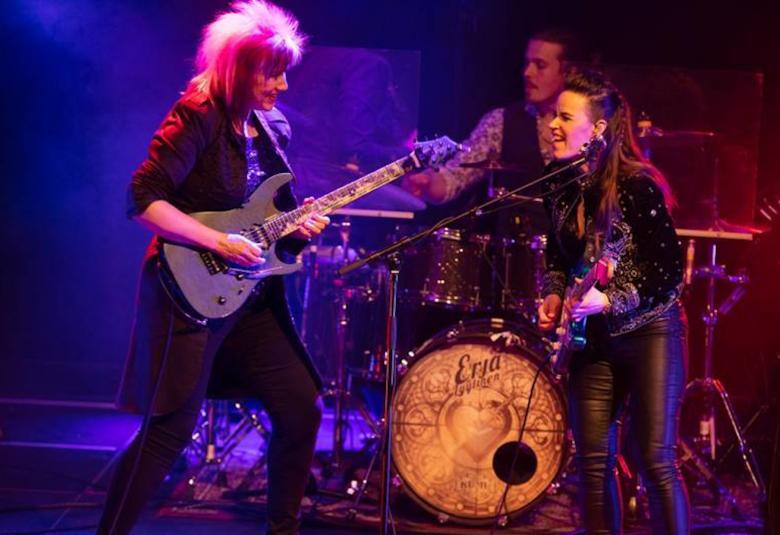 Kuvassa ovat esiintymislavalla Jennifer Batten ja Erja Lyytinen kitaroineen nahka-asuissa.  Tausta on on valaistu sinisellä ja punertavalla.