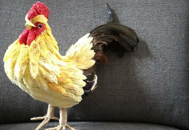 Kuvassa on keltainen pääsiäiskukko, jolla on tumma pyrstö.  Kukko on harmaalla istuimella.