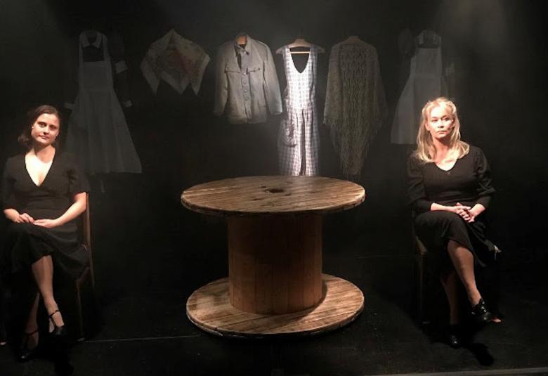 Kuvassa on tummalla näyttämöllä ja keskellä on iso pöytä.  Näyttelijät Pihla Maalismaa ja Kirsi Tarvainen ovat eri puolilla pöytää.