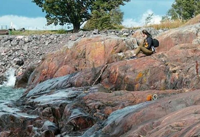 Kuvassa on Vallisaaren kallio kesäpäivänä ja alla on vettä. Kalliolla istuu yksi nainen.