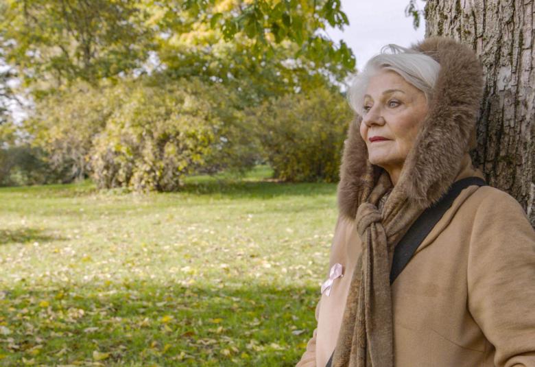 Kuvassa on Ritva Oksanen, joka nojaa puuhun hupullisessa takissa ja katsoo puistomaisemaa.