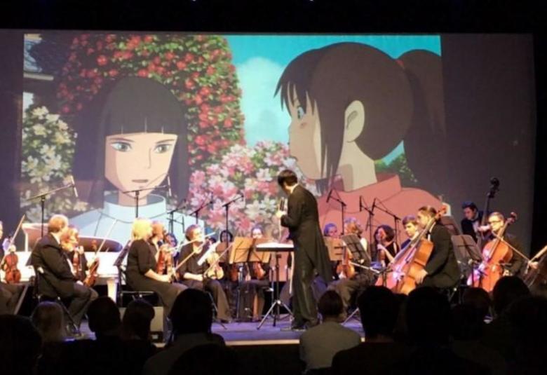Kuvassa on St Petersburg Symphony Orchestra Anime-konsertissaan.  Taustalla näkyy värikkäitä Anime-hahmoja.