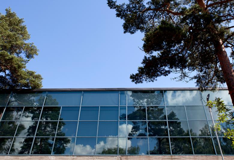 Kuvassa on EMMA taidemuseon lasinen julkisivu yläviistosti.  Taustalla on sininen taivas ja mäntyjä.