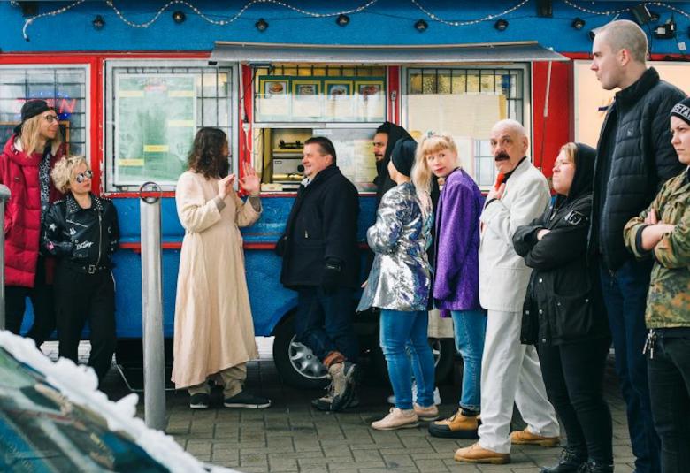 Kuvassa on ihmisiä värikkään nakkikioskin jonossa. Jonossa ovat myös Jeesus, joka katselee jonoa ja Muhammed, joka jonottaa.