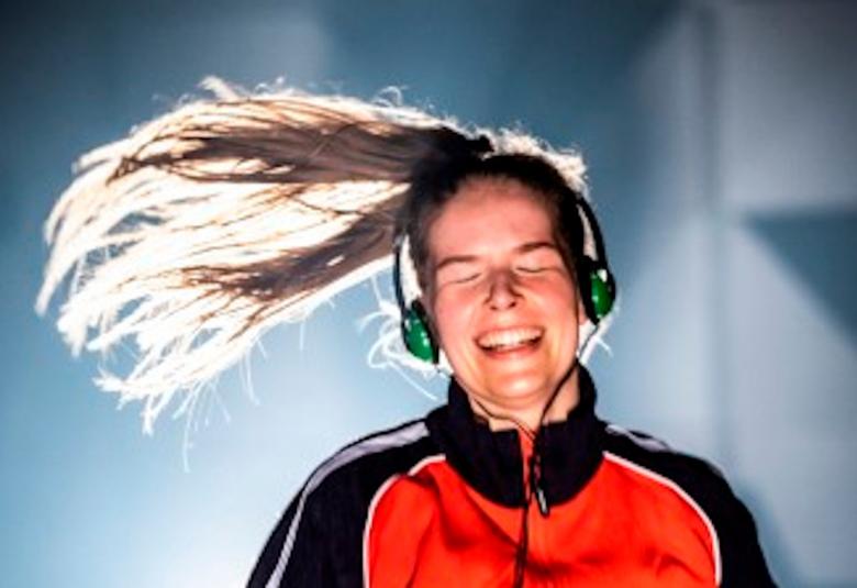 Kuvassa on näyttelijä Noora Koski kuulokkeet korvissaan ja pitkät hiukset hulmuavat vasemmalle.  Koskella on punamusta verryttelypusero päällään.