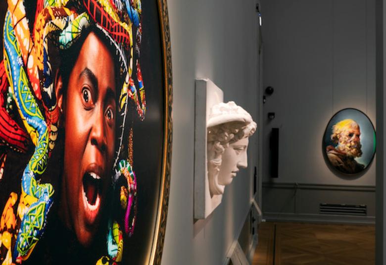 Kuvassa on vasemmalla oleva suuri taulu, jossa on tumman miehen kasvot ja päässä värikkäitä kiehkuroita.  Taaempana on pieniä tauluja ja kohokuva.