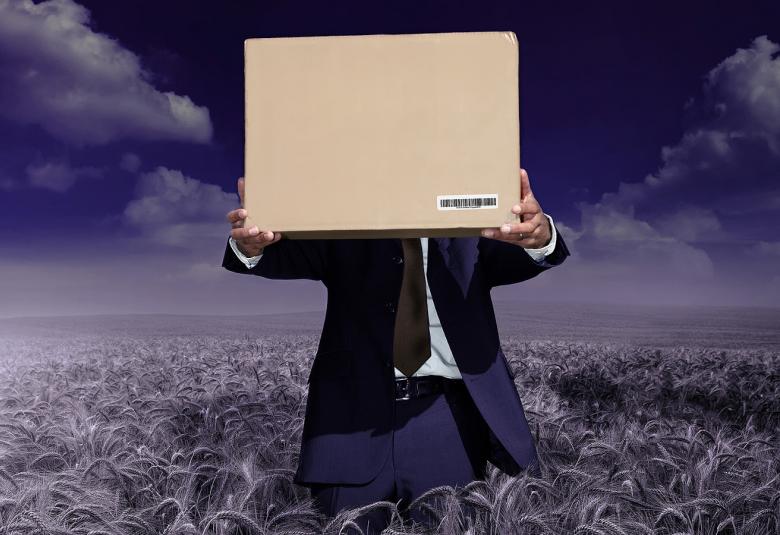 Kuvassa on mies, joka pitää tietokonetta kasvojensa edessä ja seisoo heinäpellossa jalkojaan myöten.  Kuvan sävy on tummahko.