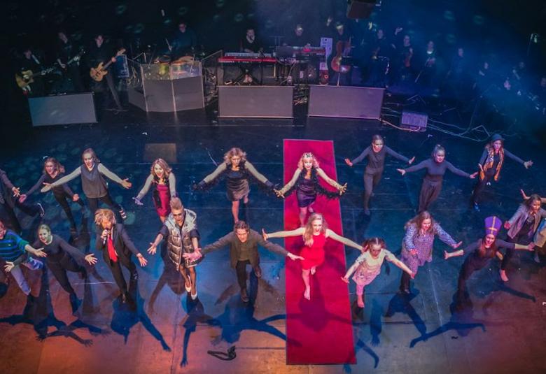 Kuvassa on nuoria näyttelijöitä esiintymässä suurena ryhmänä kahdessa rivissä ja kädet sivusuunnassa ojennettuna.  Takana on pieni orkesteri.  Kuvassa on sinistä valaistusta.