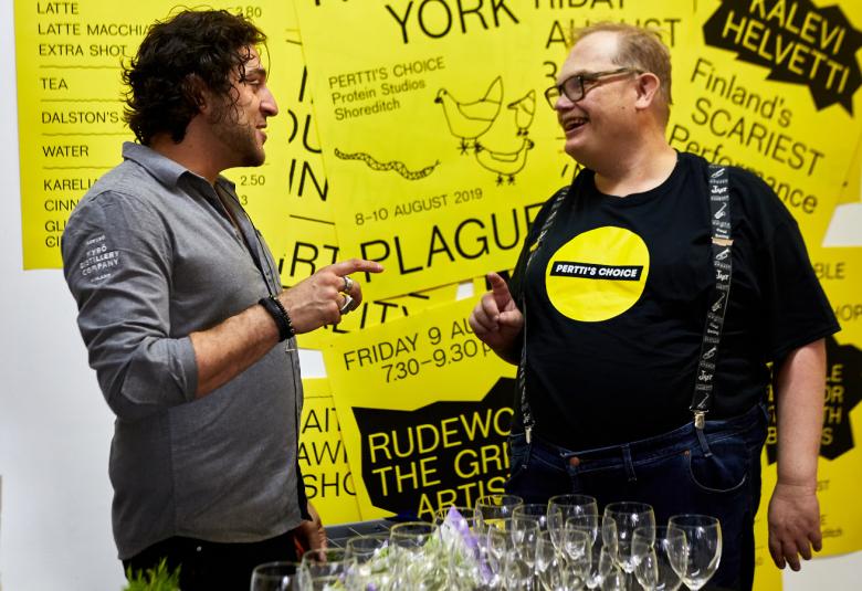 Kuvassa on Pertin Valinnan Sami Helle oikealla ja Kyrö Distilleryn edustaja kesksutelemassa edessään pöytä, jossa on laseja. Takana on keltaisella pohjalla julisteita mustalla tekstillä.