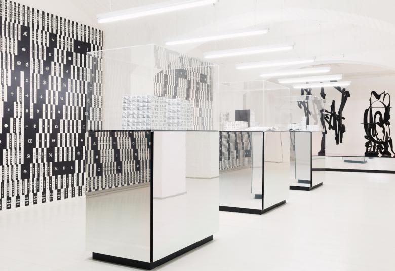 Kuvassa on näkymä Schick Toikan näyttelyhuoneesta, jossa on vitriinejä ja teoksia seinillä. Näkymä on musta-valkoinen.