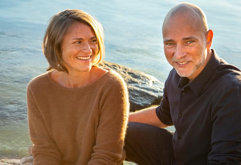 Kuvassa ovat Inka Kallen ja Aki Louhimies istumassa auringoisella ilmalla rantakalliolla meren rannalla.