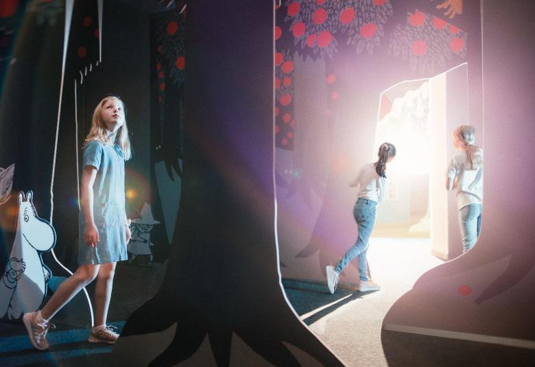 Kuvassa on tyttöjä kävelemässä muuminäyttelyssä, jossa on kankaita muumimailmakuvilla.