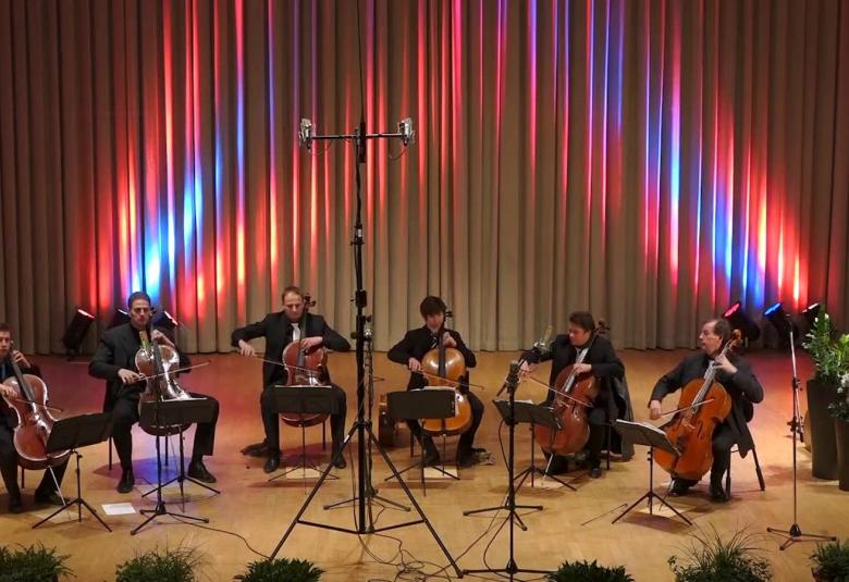 Kuvassa Total Cello Enseble soittamassa näyttämöllä, jossa on sinertävä valaistus ja tila on ruskea.