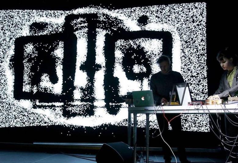 Kuvassa on seinälle heijastettu taideteos, jossa on mustat ruutumaiset kuviot keskellä. Oikealla on kaksi henkilöä pöydän ääressä läppäreiden ääressä.  