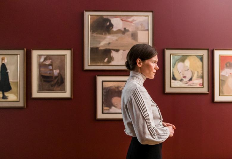 Kuvassa on Laura Birn Helenenä sivukuvassa puna-ruskealla seinällä olevien taulujen edessä. 