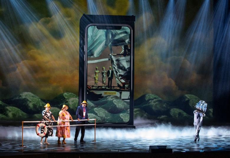 Kuvassa on oopperan näyttämöllä oikealla seniori-ikäiset kaksi naista ja yksi mies.  Keskellä on kännykän muotoinen iso taulu, jossa on heijastuskuva.  Näyttämö on tummahko ja siinä on valospotteja.