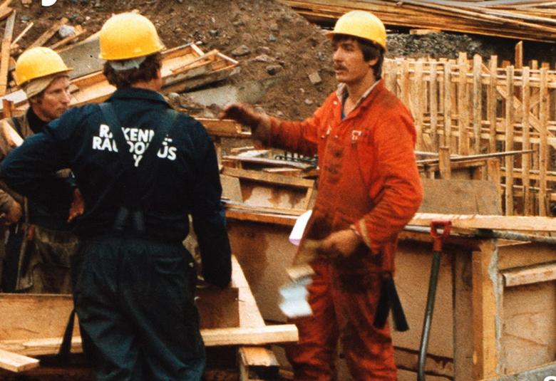 Kuvassa on rakennustyömiehiä oransseissa kypärissään rakennustyömaalla.