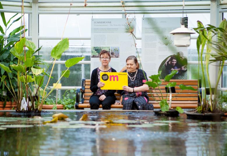 Kuvassa on kaksi naista istumassa iso museokortti kädessään ja he istuvat vesialtaan äärellä. Altaan vieressä on viherkasveja.