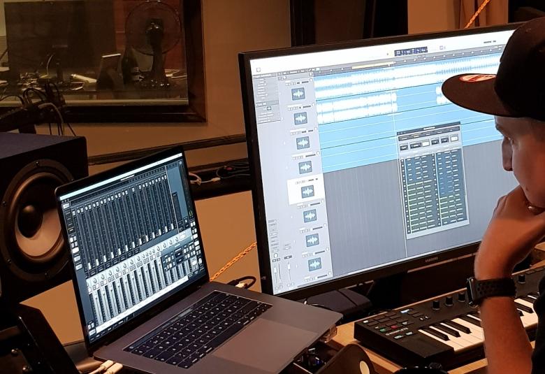 Kuvassa on Olli Saksa studiossaan, jossa näkyy Olli Saksa lippalakissa oikealla ja studiopöytä sekä läppäri edessään.