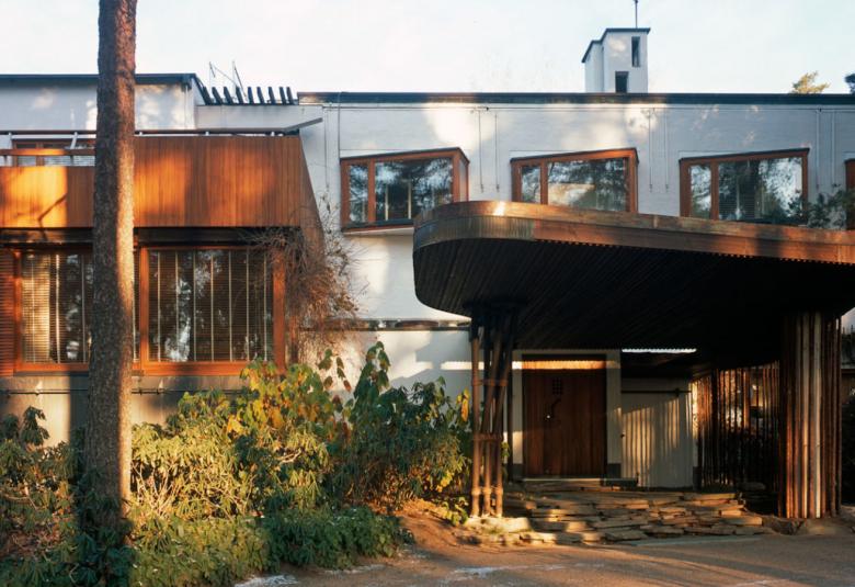 Kuvassa on Villa Mairean edusta, jossa on kaareva lippa edustan portaiden päällä ja näkyy taloa sekä kasvillisuutta.