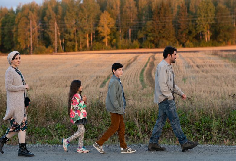 Kuvassa on Mehdipourin perhe kävelemässä peräkkäin maantien laitaa ja taustalla on keltainen viljapelto sekä kauempana metsää.