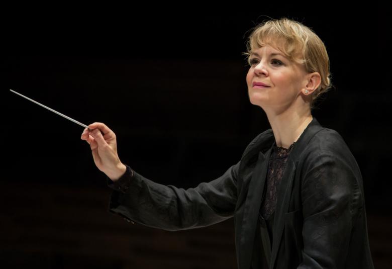 Kuvassa on Susanna Mälkki johtamassa orkesteria.  Kuva on puolivartalokuva ja Mälkillä on tahtipuikko kädessään.